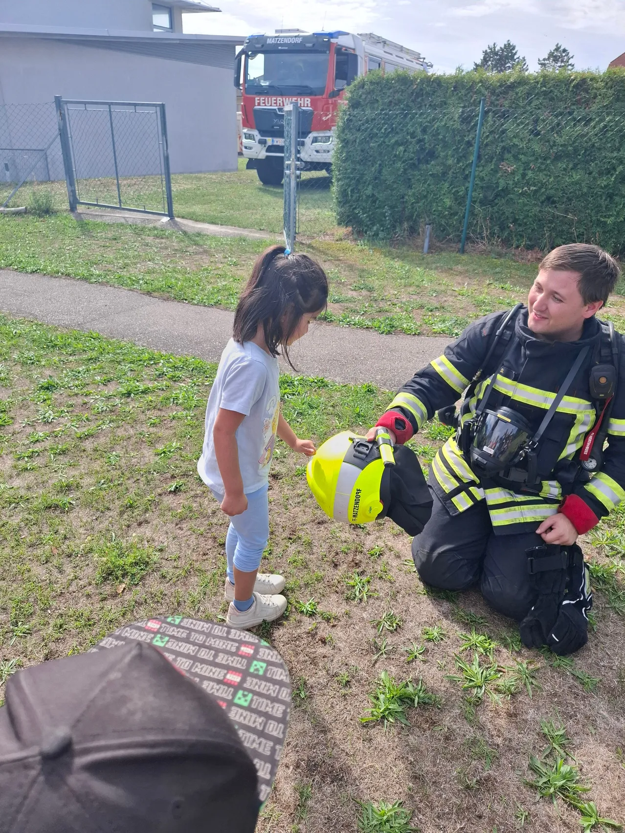 Ein Feuerwehrmann kniet auf dem Rasen und zeigt einem Kind einen gelben Helm. Das Kind schaut neugierig auf den Helm. Dahinter steht ein Feuerwehrwagen in der Nähe eines Zauns.
