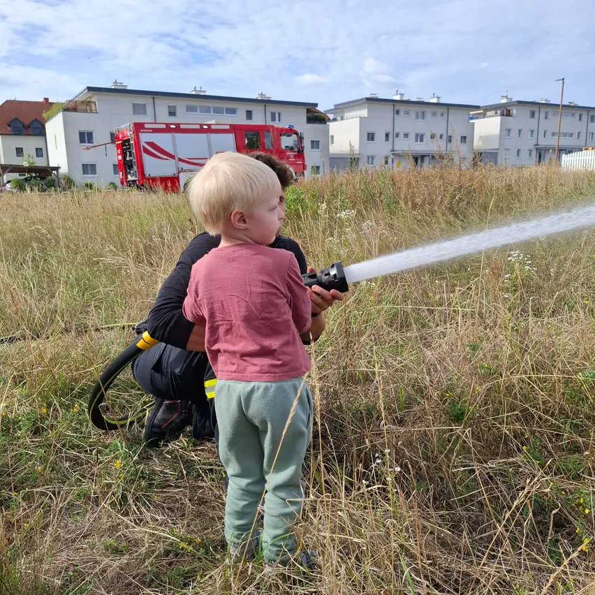 Ein junges Kind hält einen Feuerwehrschlauch, während ein Feuerwehrmann hinter ihm kniet und Wasser in einem Feld mit hohem Gras und Gebäuden im Hintergrund sprüht.
