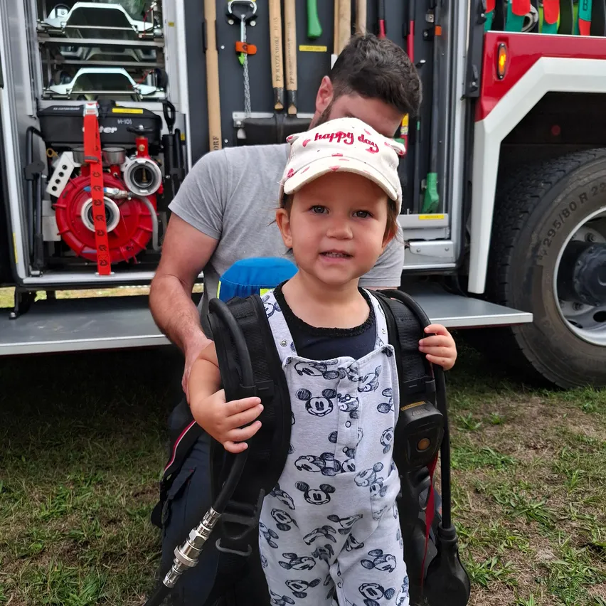 Ein kleines Kind in einem Micky-Maus-Outfit und einem Feuerwehrhelm steht vor einem Feuerwehrauto, mit einem Feuerwehrmann, der hinter ihm kniet.