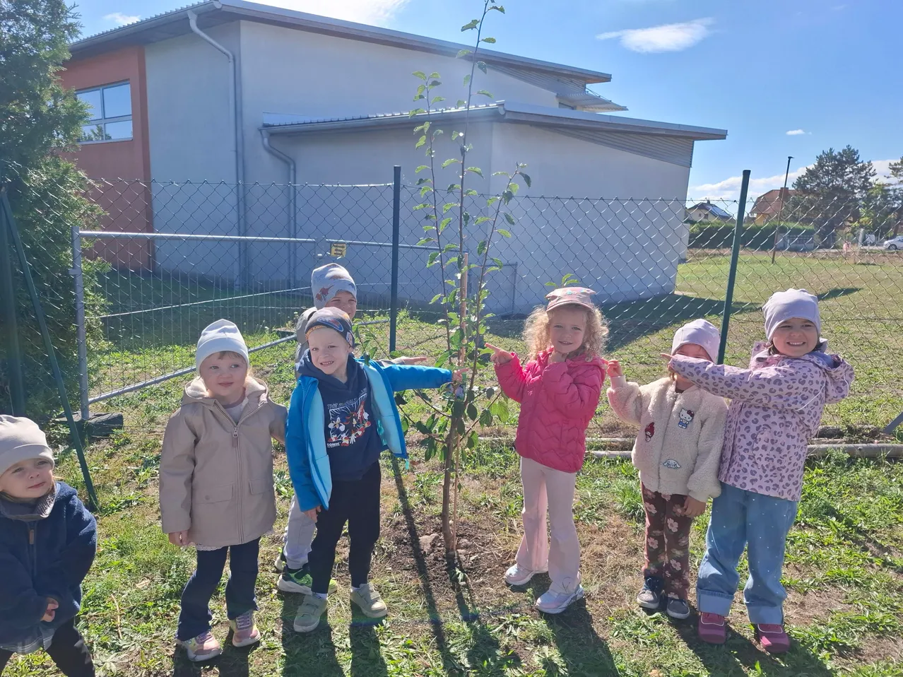 Eine Gruppe von Kindern steht um einen neu gepflanzten Baum im Garten. Sie alle lächeln und einige berühren den Baum. Hinter ihnen steht ein weißes Gebäude mit einem Zaun.