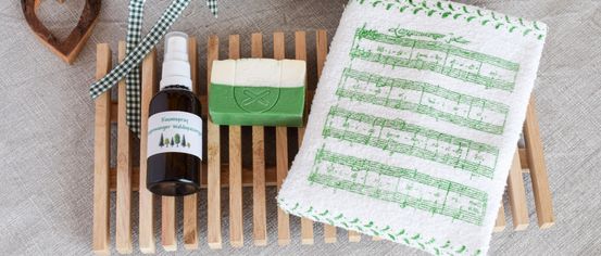 Ein Geschenkset mit einem grün gemusterten Handtuch, einer Seife und einer Flasche Raumspray, alles auf einem Holzseifenablage angeordnet.