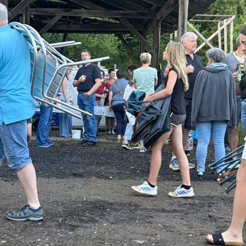 A group of people are gathered in an outdoor area, some are carrying bags and wearing sneakers. A man is holding a sandwich and standing near a table. The area is surrounded by a wooden structure.