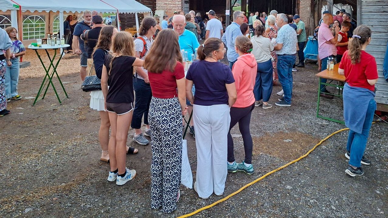 A group of people stand in a queue outdoors, some holding bottles, with a yellow hose on the ground and a tent in the background.