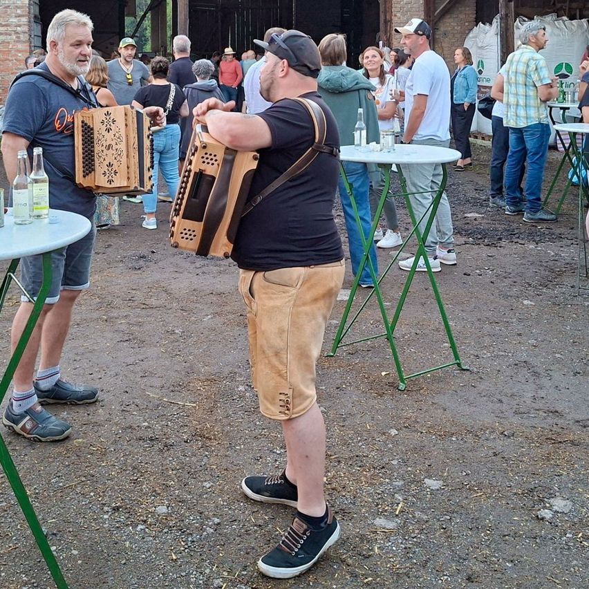A group of people are gathered outside, some playing instruments. A man with a backpack plays an accordion while another man in a hat plays a guitar.