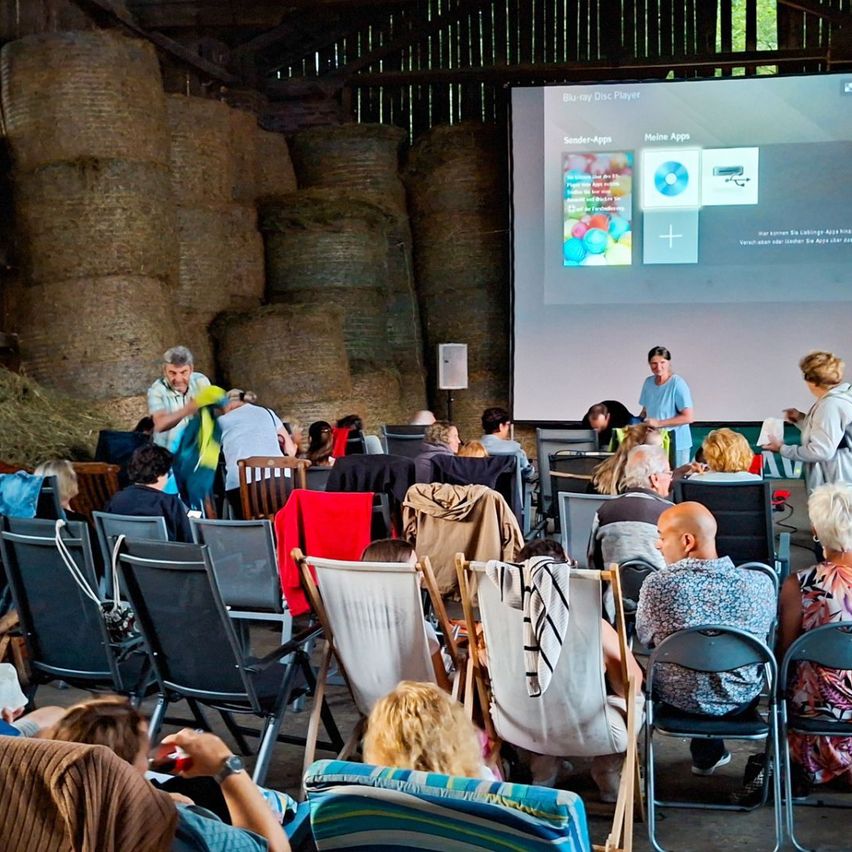 A group of people are seated in chairs in a barn, watching a presentation on a projector screen. The screen shows a Blu-ray Disc Player interface.