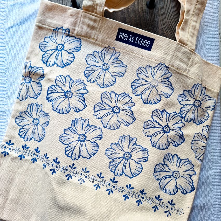 Ein beigefarbener Tragetasche mit blauem Blumendruck und einem Etikett, das 'mehr so sche' steht. Die Tasche hat einen hölzernen Hintergrund.