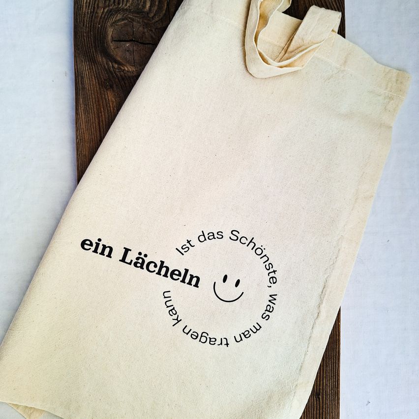 Ein beigefarbener Tragetasche mit einem lächelnden Gesicht und dem Text 'ein Lacheln' darauf gedruckt. Die Tasche ist gefaltet und auf einer Holzoberfläche platziert.