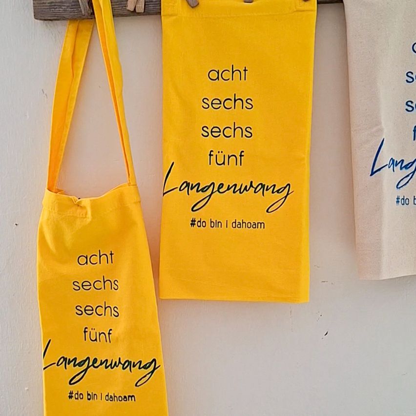 Drei gelbe Stofftaschen mit dem Text 'acht sechs sechs fünf' und 'Langenwang' hängen an einer weißen Wand. Die Taschen haben einen gelben Griff und der Text ist in Blau geschrieben.