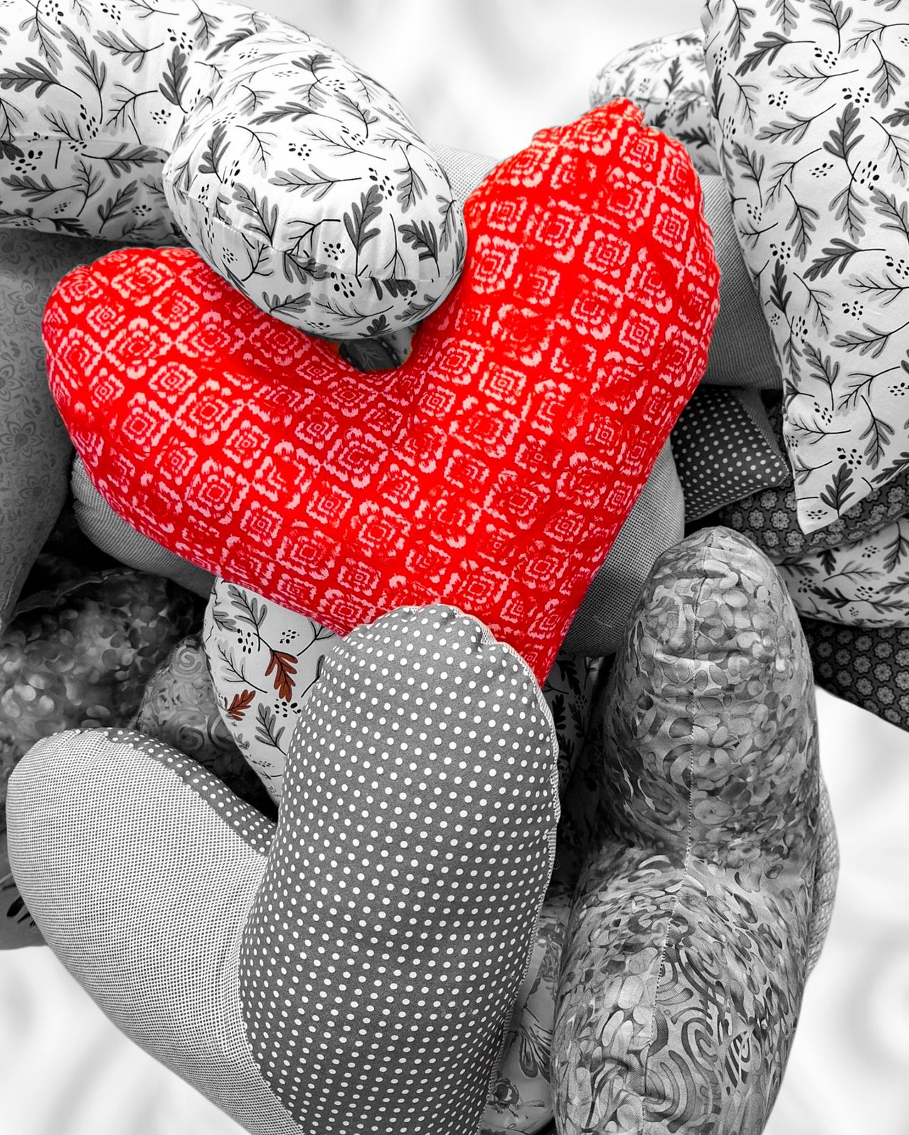 A heart-shaped pillow, red with geometric patterns, rests among various other pillows with floral and polka-dot designs. The pillows are in black and white tones.