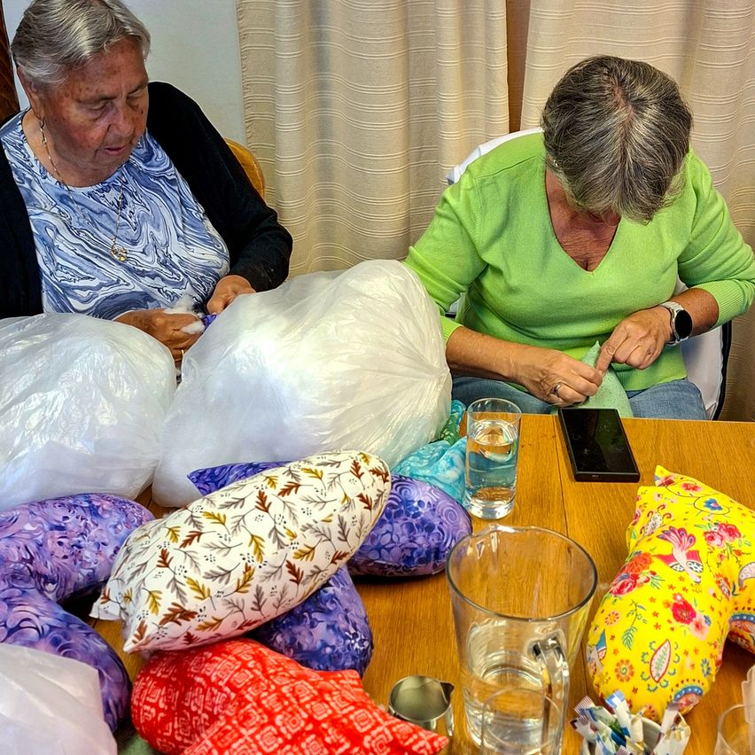Two older women sit at a table filled with pillows and bags. One woman holds a cell phone, and both have items in their hands. A glass of water and a phone are on the table.