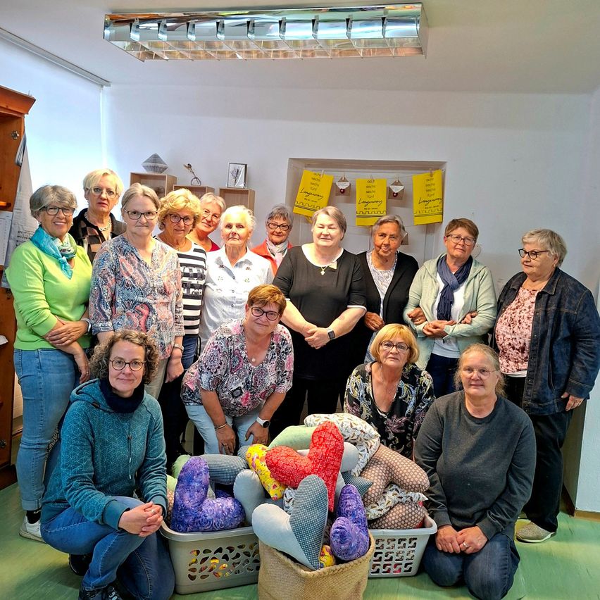 A group of women are posing for a photo in a room with several yellow papers on the wall. Some are sitting on the floor while others are standing. They are all smiling and holding some stuffed toys.