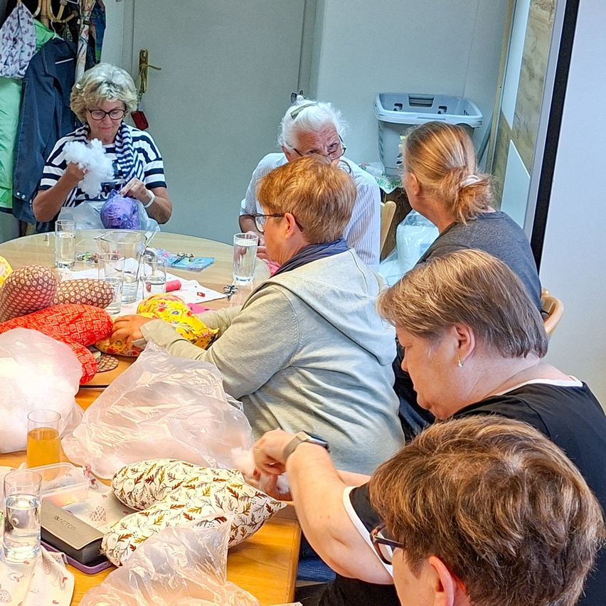 Several women are gathered around a table in a room. They are engaged in various activities such as sewing and using a knitting machine. The table is filled with fabric, pillows, and other craft materials. There are clothes hanging on the wall in the background.