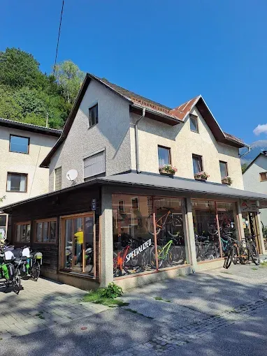 Ein Fahrradgeschäft mit vielen ausgestellten Fahrrädern an der Vorderseite. Im Hintergrund sind Bäume und ein Haus zu sehen.