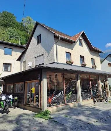 Ein Fahrradgeschäft mit vielen ausgestellten Fahrrädern an der Vorderseite. Im Hintergrund sind Bäume und ein Haus zu sehen.