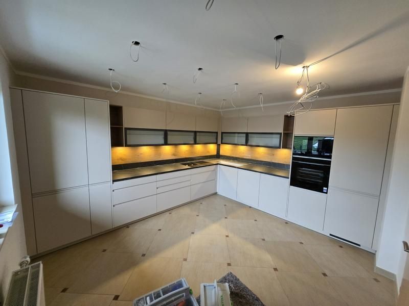 Eine leere moderne Küche mit weißen Schränken, einem Holzboden und einer schwarzen Arbeitsplatte. Die Küche ist durch Deckenlampen beleuchtet.