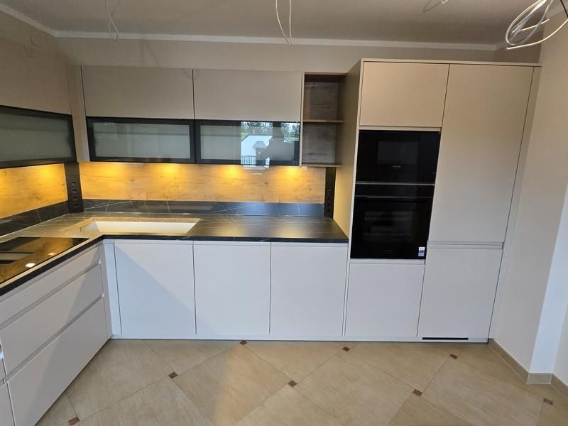 Eine moderne Küche mit weißen Schränken, einer schwarzen Marmor-Arbeitsplatte und eingebauten Geräten. Die Küche verfügt über ein Waschbecken, eine Mikrowelle und einen Backofen. Der Raum hat einen Holzboden und ein Glasfenster.