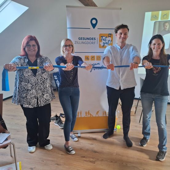 Vier Erwachsene stehen mit einem blauen Band in der Hand und lächeln in einem Raum mit Holzboden und einer weißen Wand mit einem Poster für 'Gesundes Zillingdorf'.