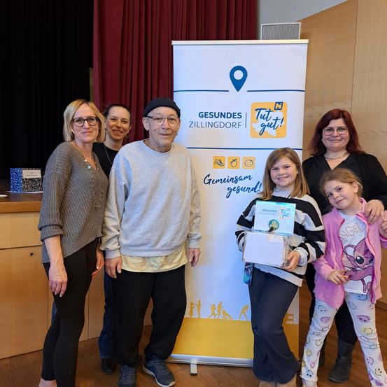 Eine Gruppe von Menschen, darunter zwei junge Mädchen, ein älterer Mann und drei Frauen, stehen vor einem Banner mit den Worten 'Gesundheit Zillingdorf' und 'Tut gut!' Sie lächeln und posieren für ein Foto.