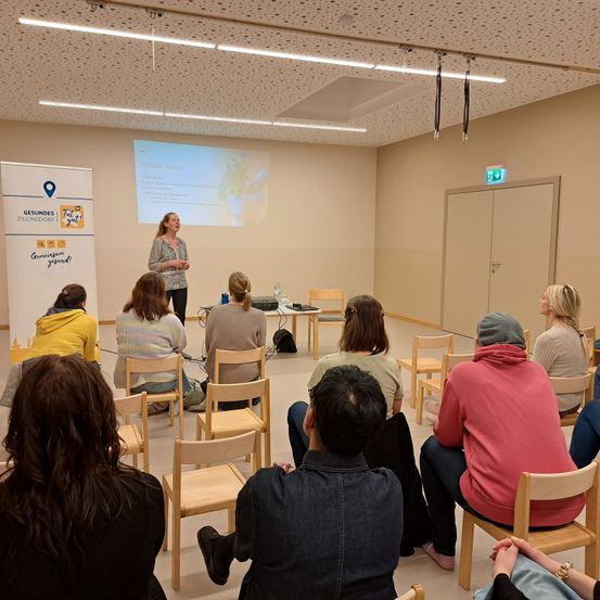 Eine Frau steht und spricht zu einer Gruppe in einem Raum mit Stühlen, einer Projektionsleinwand und einem Banner. Der Raum hat eine beige Wand und Decke mit Leuchten.