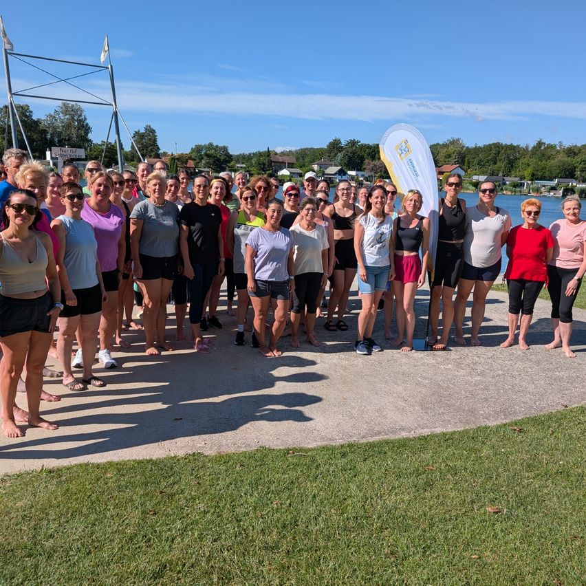 Eine Gruppe von Frauen in Badekleidung und Freizeitkleidung posiert für ein Foto am See. Sie stehen auf Beton mit Gras und Bäumen im Hintergrund.