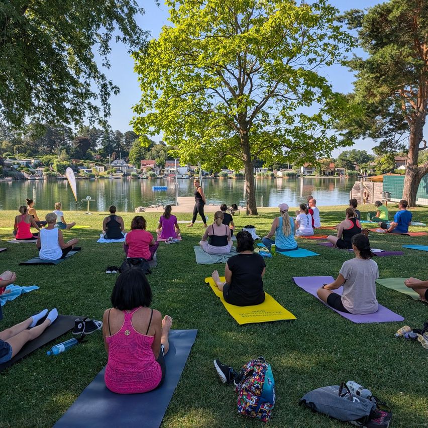 Eine Gruppe von Menschen praktiziert Yoga auf dem Rasen in der Nähe eines Sees. Eine Frau leitet die Yoga-Sitzung.