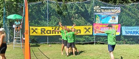 Eine Gruppe von Menschen spielt Volleyball auf einem Rasenfeld mit Netz und Schiedsrichter. Ein Mann steht unter einem Schirm, und es gibt ein Banner mit dem Text Raiffeisen und ein weiteres mit dem Wort SPARK.