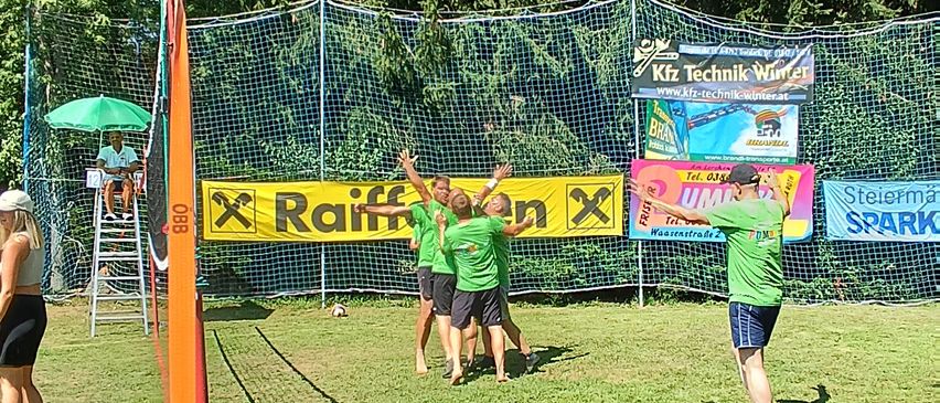 A group of people are playing volleyball on a grassy field with a net and a referee. A man stands under an umbrella, and there is a banner with the text Raiffeisen and another with the word SPARK.