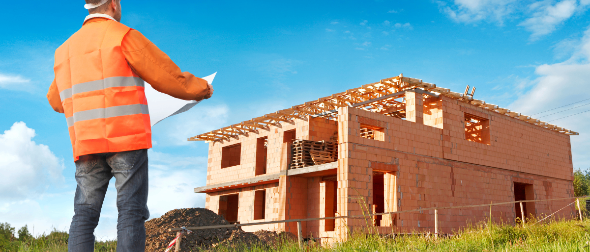 Ein Bauarbeiter hält einen Bauplan und sieht ein Haus im Bau mit Ziegelwänden und Holzdach.