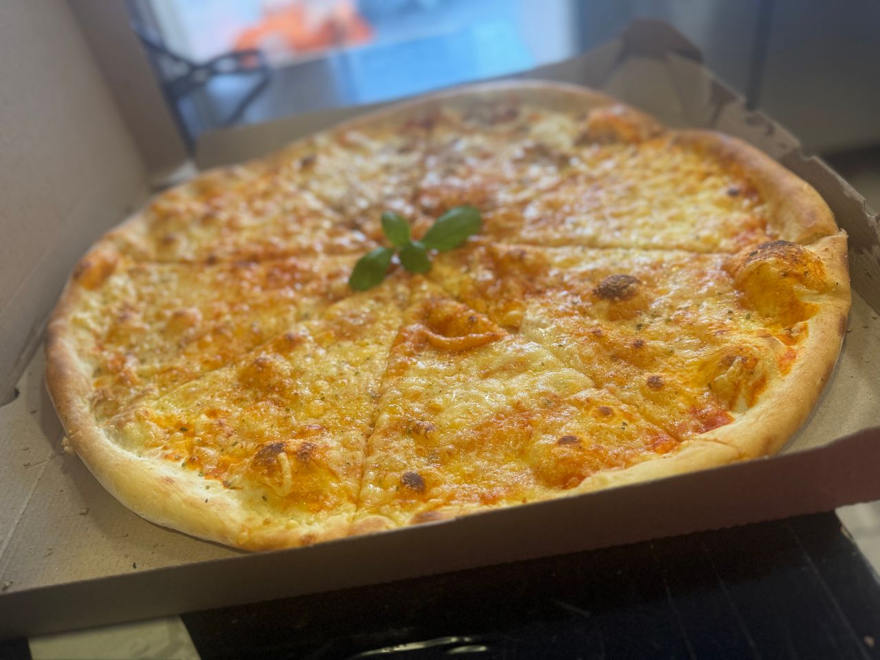 Eine ganze Pizza mit geschmolzenem Käse und einem Basilikumblatt in einer Pappschachtel auf einem Tisch.