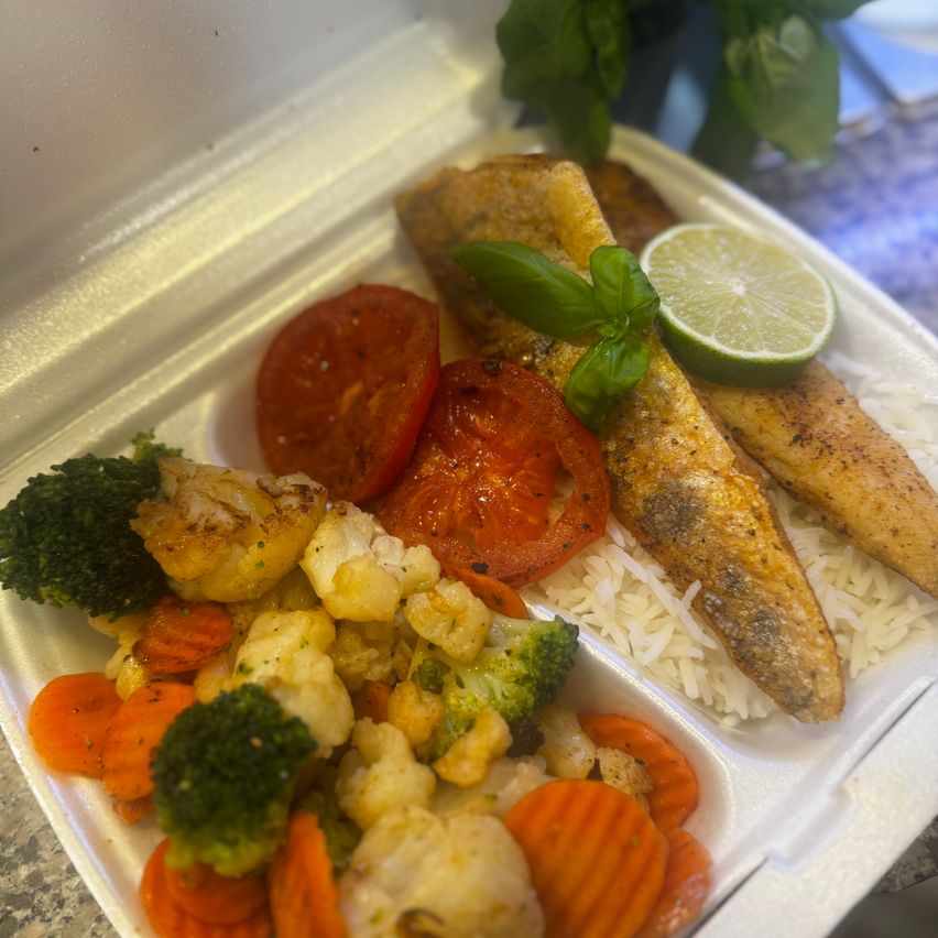 Ein rechteckiger weißer Behälter enthält eine Mahlzeit mit gegrilltem Fisch, Reis, Brokkoli, Blumenkohl, Karotten und Tomaten. Eine Limettenscheibe und ein Basilikumblatt schmücken den Fisch.