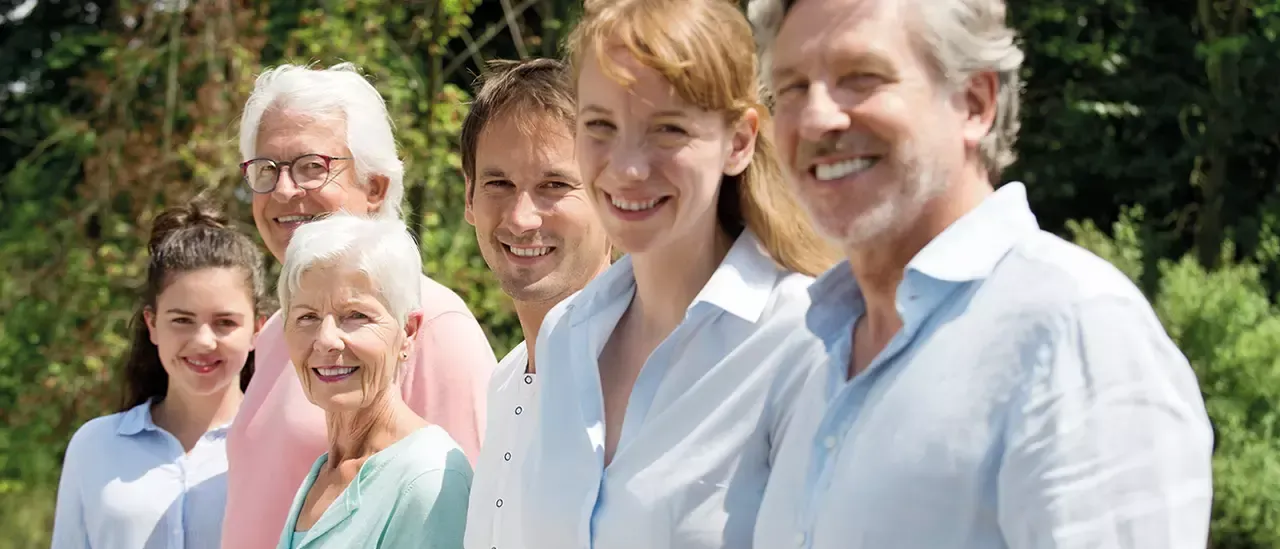 Fünf lächelnde Personen, zwei Männer und drei Frauen, stehen vor einem üppigen grünen Hintergrund. Sie tragen alle helle Hemden, und die Frauen tragen Blusen.