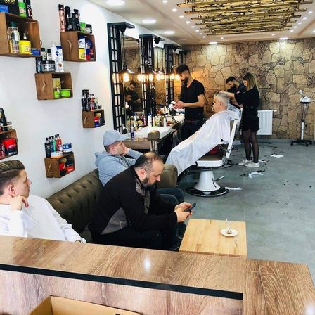 Mehrere Männer befinden sich in einem Friseurladen. Ein Mann lässt sich die Haare schneiden. Ein anderer Mann benutzt sein Handy. Der Laden hat eine Holztheke, eine Couch und Regale mit Haarpflegeprodukten.