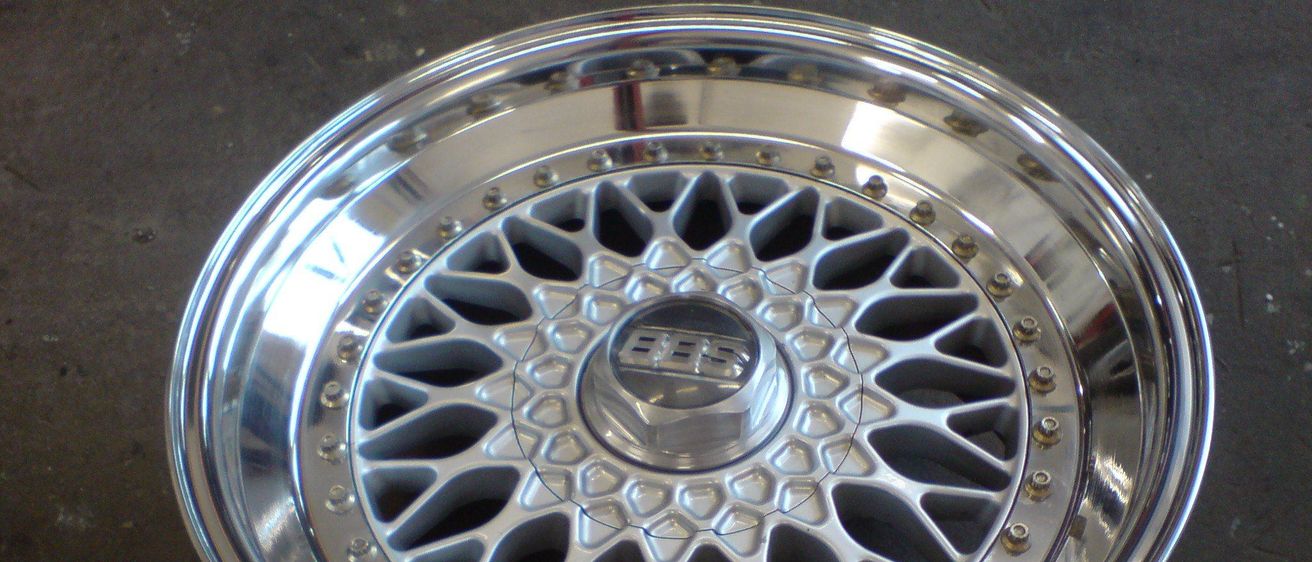 Das Bild zeigt eine Nahaufnahme eines polierten silbernen Leichtmetallrads mit komplizierten Speichen und einer zentralen Radkappe. Der Felgenrand hat eine glatte Oberfläche, und die Speichen weisen ein Diamantmuster auf.