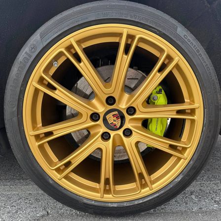 Eine Nahaufnahme eines schwarzen Autorads mit goldenen Speichen und einem gelben Bremssattel. Die Felge zeigt das Porsche-Logo.