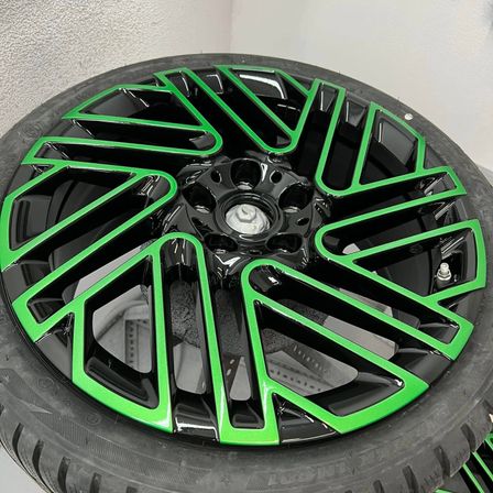 Ein schwarzer Reifen mit einer glänzenden grünen Felge ist auf einem schwarzen Reifen montiert. Der Reifen hat eine silberne Radkappe. Die Felge ist poliert und hat ein sternförmiges Design. Der Reifen ist auf einer weißen Oberfläche platziert.