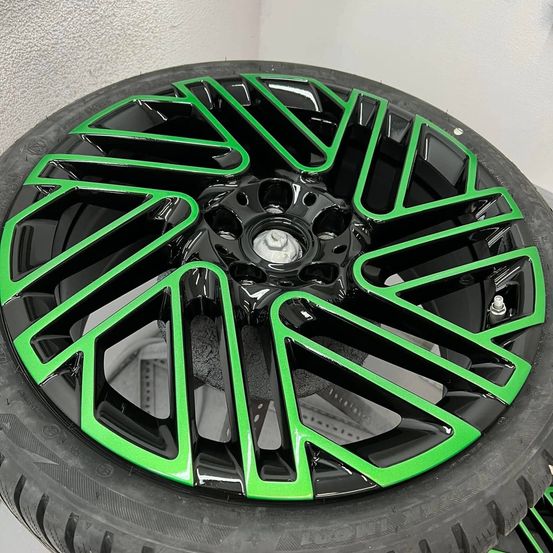 Ein schwarzer Reifen mit einer glänzenden grünen Felge ist auf einem schwarzen Reifen montiert. Der Reifen hat eine silberne Radkappe. Die Felge ist poliert und hat ein sternförmiges Design. Der Reifen ist auf einer weißen Oberfläche platziert.