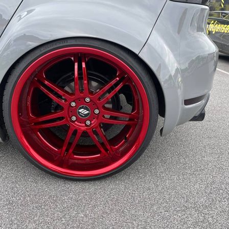 Nahaufnahme des hinteren Rades eines grauen Autos mit roten Leichtmetallfelgen und schwarzen Reifen. Das Auto steht auf einem Parkplatz.