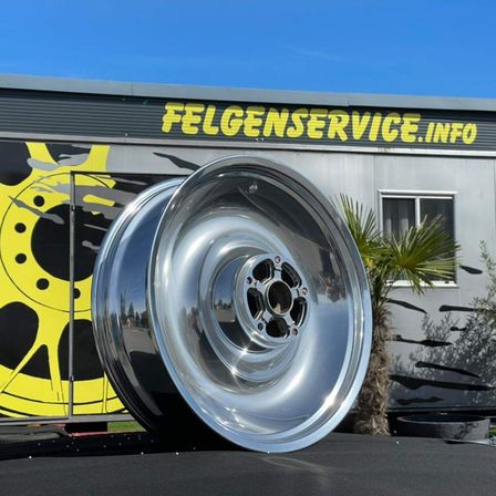 Ein glänzendes Chromrad wird vor einem Gebäude mit dem Text Felgenservice.info an der Wand ausgestellt. Ein gelbes Raddesign befindet sich auf der linken Seite des Gebäudes.