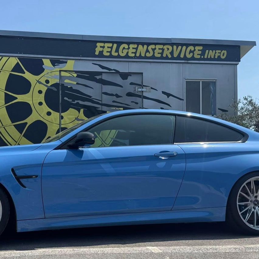 Ein blauer Sportwagen steht vor einem Gebäude mit dem Schild 'FELGENSERVICE.INFO'. Der Wagen ist glänzend und hat einen gelben Felgenrand. Hinter dem Auto steht ein Gebäude mit einem großen Plakat eines Rades.