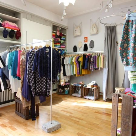 Ein Bekleidungsgeschäft zeigt eine Vielzahl von Kleidungsstücken an Kleiderständern, mit Regalen gefüllt mit Taschen und Hüten. Accessoires hängen an der Wand, und Holzkisten stehen auf dem Boden. Der Boden ist aus Holz, und die Wände sind weiß.