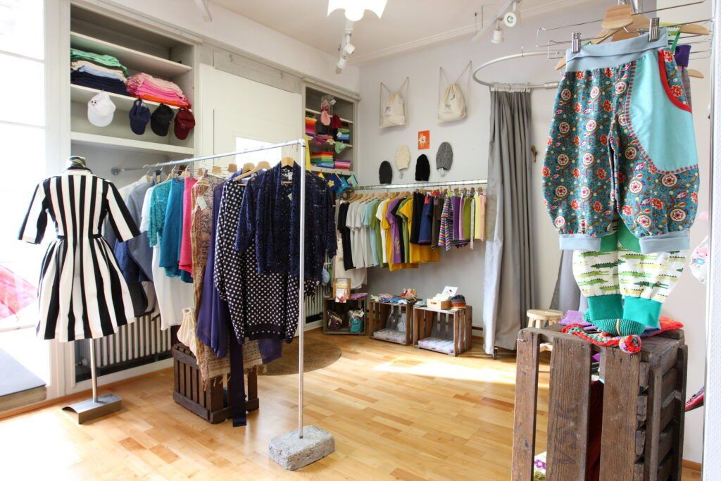 Ein Bekleidungsgeschäft zeigt eine Vielzahl von Kleidungsstücken an Kleiderständern, mit Regalen gefüllt mit Taschen und Hüten. Accessoires hängen an der Wand, und Holzkisten stehen auf dem Boden. Der Boden ist aus Holz, und die Wände sind weiß.