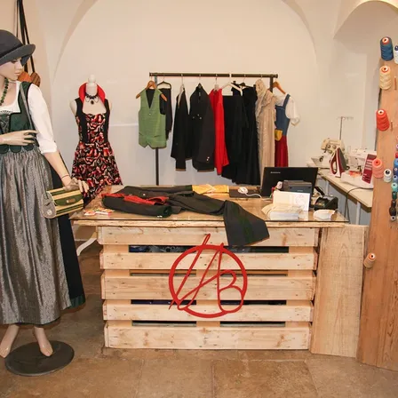 Ein Boutique mit verschiedenen Kleidern auf einem Holzständer. Ein Mannequin in traditioneller Kleidung steht in der Nähe. Der Tresen hat das AB-Logo.