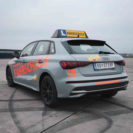 Ein grauer Audi A3 mit einem gelben Schild auf dem Dach, das 'Fahrschule' steht. Das Auto ist auf einem Betonuntergrund mit Reifenspuren geparkt.