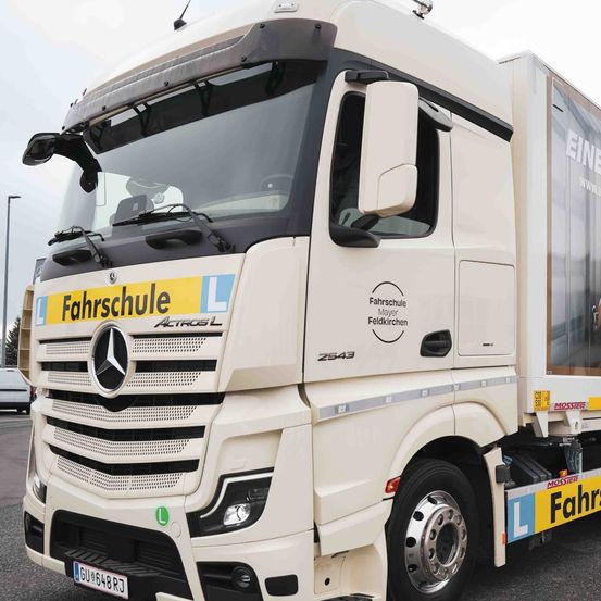 Ein weißer Mercedes-Benz Actros LKW mit gelben und blauen Aufklebern ist auf einem Parkplatz geparkt. Die Stoßstange vorne hat ein gelbes Schild mit der Aufschrift 'Fahrschule'. Der LKW hat eine große Seitenwerbung.