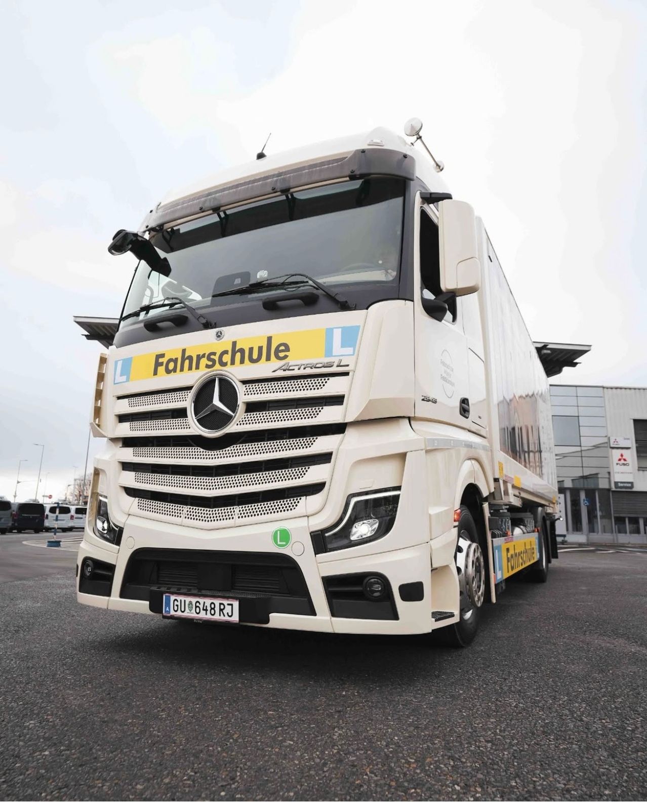 Ein weißer Mercedes-Benz Actros Lkw mit einem gelben Schild, das 'Fahrschule' steht, ist in einem Parkplatz geparkt, mit Gebäuden und geparkten Autos im Hintergrund.