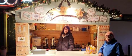 Eine Frau steht lächelnd hinter einem festlich geschmückten Tresen. Der Tresen ist mit Weihnachtslichtern verziert, und verschiedene Gegenstände wie Flaschen und Behälter sind ausgestellt. Hinter ihr befinden sich Kaffeeautomaten und ein kleiner Herd.