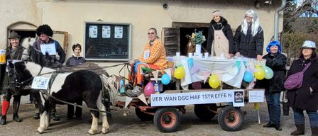 Eine Gruppe von Menschen in Kostümen steht neben einer Pferdekutsche vor einem Gebäude. Ein Banner steht: VER WAN DSC HAF TSTR EFFE.