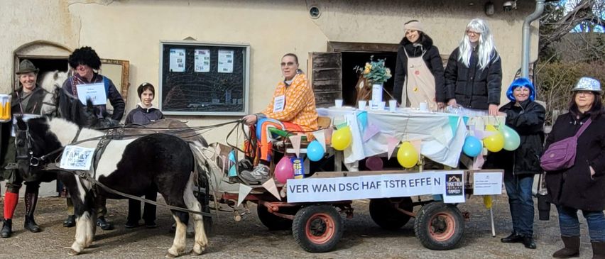 Eine Gruppe von Menschen in Kostümen steht neben einer Pferdekutsche vor einem Gebäude. Ein Banner steht: VER WAN DSC HAF TSTR EFFE.