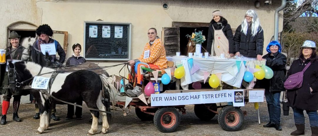 Eine Gruppe von Menschen in Kostümen steht neben einer Pferdekutsche vor einem Gebäude. Ein Banner steht: VER WAN DSC HAF TSTR EFFE.