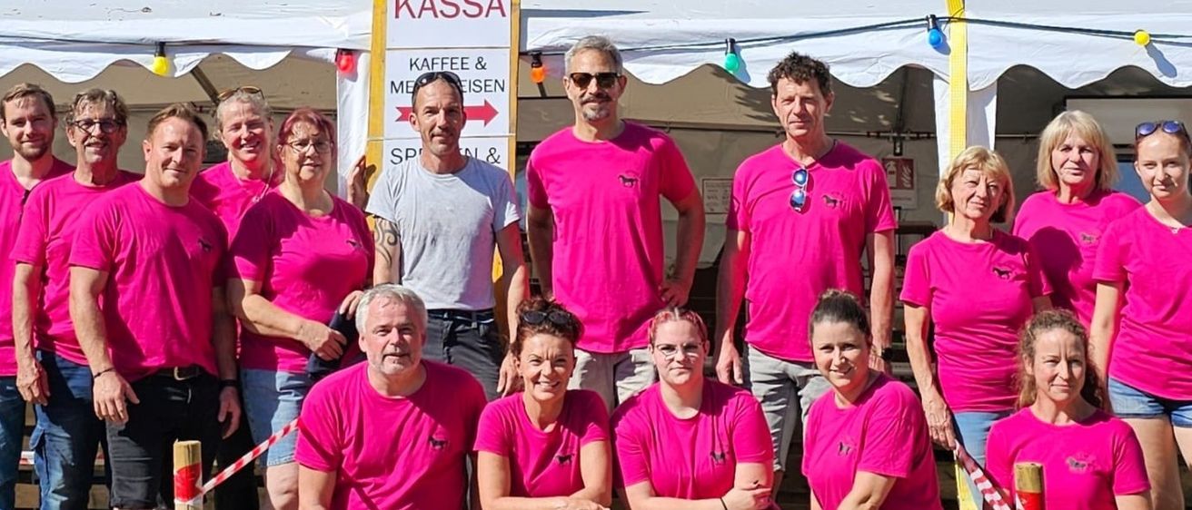 Eine Gruppe von Menschen in rosa T-Shirts posiert für ein Foto vor einem Zelt mit einem Schild, das KASSA KAFFEE & MEISIEN liest.